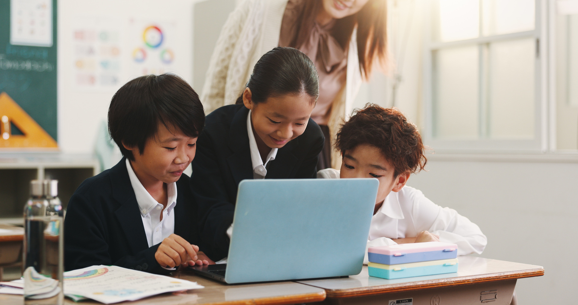 Teacher pointing at a whiteboard while delivering a lesson to students seated in a classroom Teacher providing one-to-one support to a pupil during a classroom activity at a desk Teacher supporting pupils using a laptop during a classroom lesson