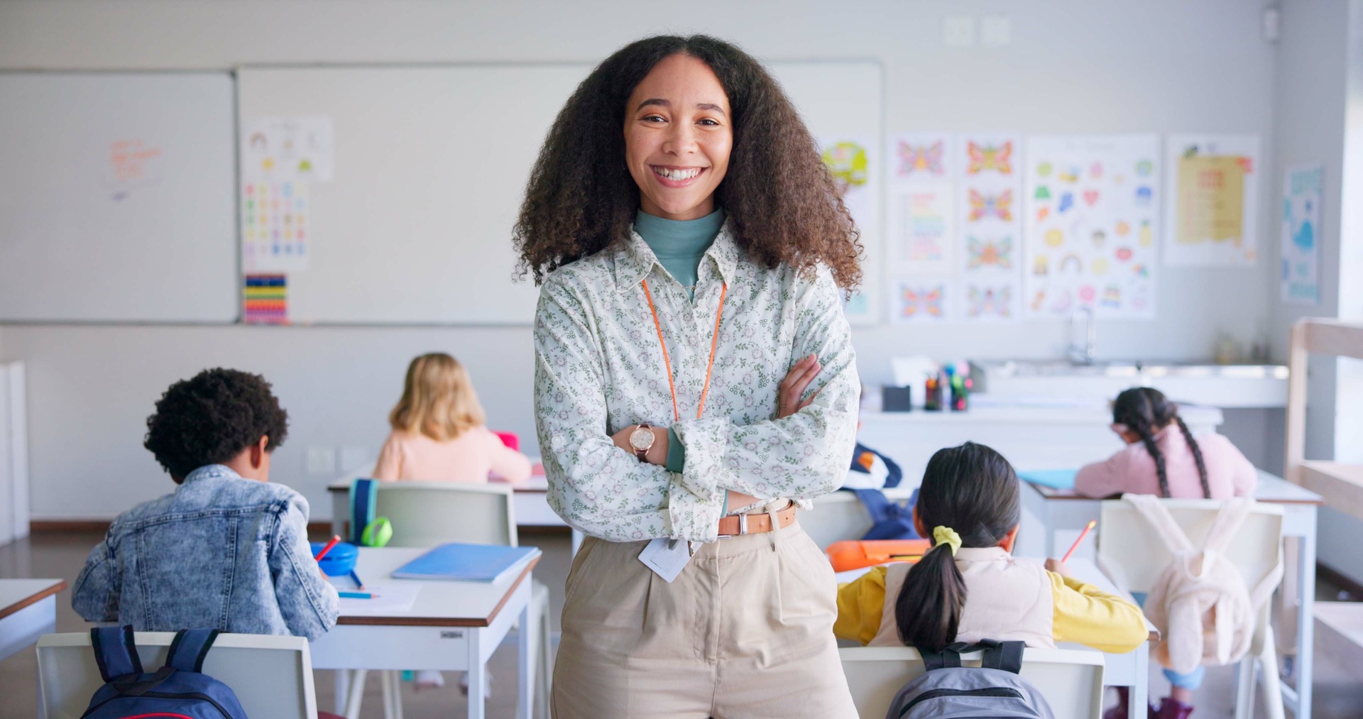 Teacher standing at the front of a classroom with arms crossed while pupils work at desks in the background